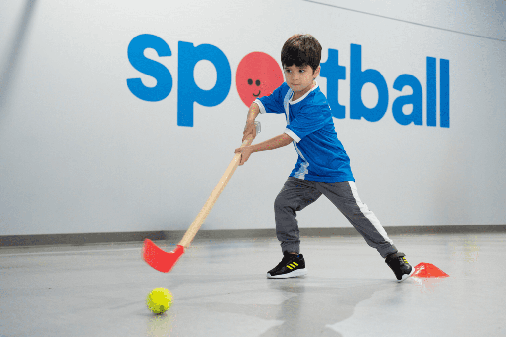 Boy practicing hockey skills in Sportball kids sports program, swinging stick at ball.